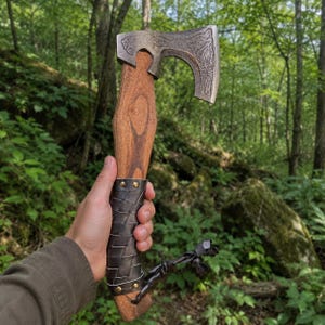 May include: A hand axe with a dark metal head featuring intricate etched patterns, a smooth brown wooden handle, and a black leather-wrapped grip secured with brass rivets. A black leather cord is tied to the base of the handle.