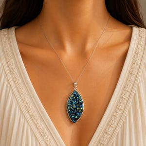 May include: A necklace featuring a marquise-shaped pendant with a textured, dark blue and iridescent crystalline surface set in a silver-toned metal frame, suspended from a thin, delicate silver-toned chain against a cream-colored fabric.