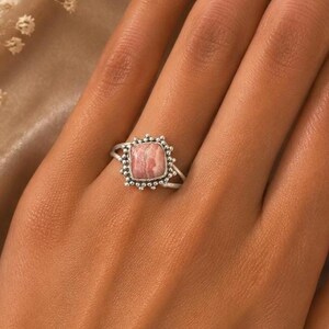May include: A silver ring featuring a square, pink and white banded stone set in a beaded metal frame. The band of the ring is split into two thin silver wires that meet at the base of the setting, creating a detailed and textured appearance.