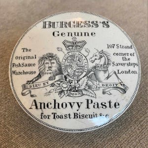 May include: A round white ceramic lid featuring black printed text and illustrations. The text reads Burgess's Genuine Anchovy Paste for Toast Biscuit &c, with a central crest depicting a lion and a unicorn flanking a crown and shield.