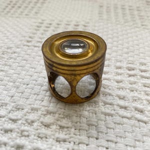 May include: A circular brass linen tester magnifying glass featuring a single glass lens set into a cylindrical frame. The base of the frame has three arched cutouts, providing a stable structure for viewing small details on flat surfaces.