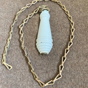 May include: A light blue, ribbed, tapered glass pull handle attached to a long, metallic, interlocking chain. The glass component features a smooth, matte finish with a rounded, tiered base and a small, dark metal cap at the top and bottom.