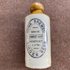 May include: A cylindrical stoneware ginger beer bottle with a tan glazed top and a cream-coloured body. The front features a black oval stamp reading HOME BREWED GINGER BEER, MAKERS FIANDER & CO, NEWPORT I. OF W. with decorative floral accents.