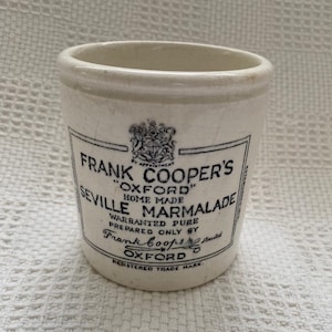 May include: A cream-coloured ceramic jar with a crackled glaze finish. The front features black printed text reading Frank Cooper's Oxford Home Made Seville Marmalade, along with a crest, warrant information, and the signature of Frank Cooper.