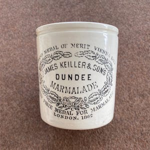 May include: A cylindrical white ceramic marmalade jar featuring black printed text that reads James Keiller & Sons Dundee Marmalade, along with Grand Medal of Merit Vienna 1873 and Prize Medal for Marmalade London 1862, surrounded by an oak leaf wreath.