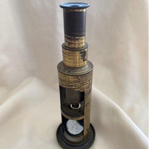 May include: A vintage brass microscope featuring a cylindrical body with a weathered, dark gold finish. The instrument includes an adjustable eyepiece, a central stage area with a circular opening, and a circular base, showing signs of age and wear.