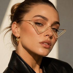 May include: Gold-toned metal glasses featuring a distinct V-shaped frame design. The thin wire rims are embellished with small, clear crystals along the outer edges. These glasses have a geometric silhouette and a polished metallic finish.