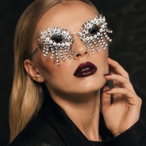 Puede incluir: Gafas decorativas con dos grandes piedras ovaladas oscuras en monturas adornadas con cristales transparentes facetados y perlas blancas. Largos flecos de cristal cuelgan de las monturas, imitando la forma de las pestañas.