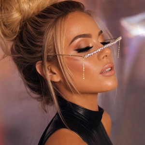 May include: A gold-toned metal eyewear accessory featuring a single horizontal bar across the bridge area, encrusted with iridescent, multi-colored crystals. A delicate chain of small, clear crystals hangs vertically from the right side.