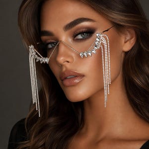 May include: Decorative eyewear featuring a thin gold-tone metal frame with a V-shaped bridge. The frame is adorned with clear, pear-shaped crystals along the top edges and long, dangling strands of sparkling rhinestones hanging from both sides.