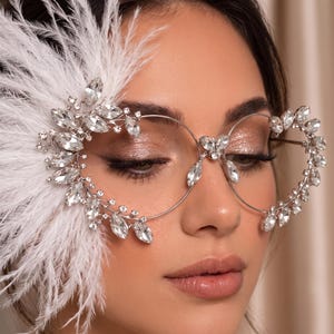 May include: Decorative eyewear featuring a thin silver-tone metal frame adorned with clear, marquise-cut crystals arranged in a floral pattern. One side of the frame is embellished with a large, white, wispy feather accent.