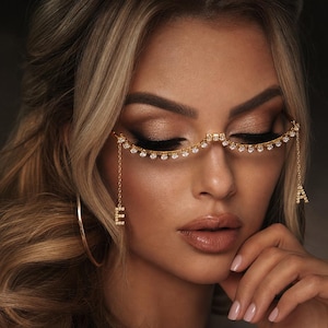 May include: A gold-toned decorative eyewear accessory featuring a curved bridge lined with clear rhinestones. Two delicate gold chains hang from the sides, each ending in a dangling letter charm, with the letter E on the left and the letter A on the right.