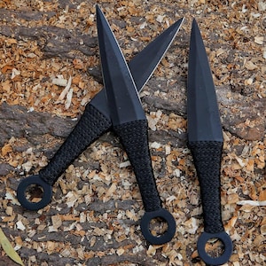Könnte beinhalten: Drei schwarze Wurfmesser mit spitzen Klingen und kordelumwickelten Griffen. Jeder Griff verfügt über einen kreisförmigen Ring an der Basis. Die Messer sind in einer Gruppe angeordnet und zeigen ihre matte Oberfläche und scharfe Metallklingen.