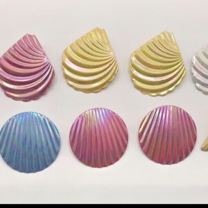 May include: A collection of twelve decorative buttons in various shapes and finishes. The set includes shell-shaped, circular, and triangular buttons featuring iridescent, metallic, and solid colors like pink, gold, silver, and blue.