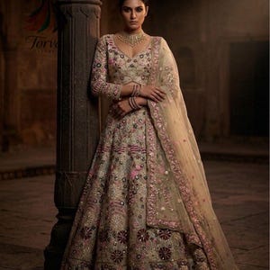May include: A beige lehenga choli featuring intricate floral embroidery in shades of pink, maroon, and blue, with metallic thread work and mirror embellishments. The ensemble includes a long-sleeved blouse, a full skirt, and a sheer dupatta.