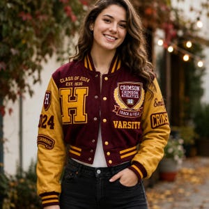 Puede incluir: Una chaqueta universitaria de color granate y dorado con la letra H, el texto Class of 2024 Est. 1636 y un emblema de Crimson Athletics Track & Field. Las mangas son doradas con puños granates y el frente incluye botones a presión dorados.