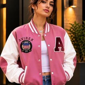 Peut inclure: Une veste universitaire rose et blanche avec une fermeture à boutons-pression blancs, un col, des poignets et un ourlet côtelés rayés. La poitrine présente un grand écusson lettre A et un écusson circulaire Avirex All American 1975.
