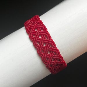 May include: A vibrant red bracelet featuring a complex, woven macrame pattern with a series of interconnected loops and knots. The design consists of a continuous, repeating lattice structure crafted from thin, durable cord, creating a textured surface.