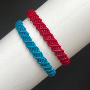 May include: Two macrame bracelets, one in a vibrant red and the other in a bright turquoise blue, featuring a consistent twisted knot pattern throughout the length of each band. The bracelets are displayed side by side on a white cylindrical form.