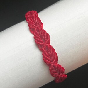 May include: A bright red macrame bracelet featuring a repeating leaf-like pattern created with intricate knotted cord. The design consists of a series of interconnected, textured segments that form a continuous, flexible band of vibrant red thread.