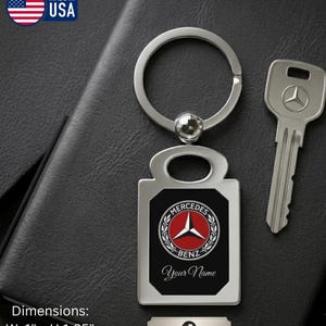 May include: A silver-toned metal keychain featuring a rectangular pendant with a black center, displaying the Mercedes-Benz logo in red, white, and black, with the text 'Your Name' below. The keychain measures 1 inch wide by 1.35 inches high.