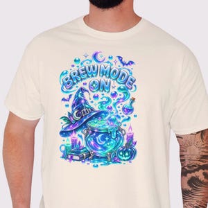 May include: A cream-colored t-shirt featuring a vibrant graphic print of a witch's hat, a bubbling cauldron, candles, a book, and a pumpkin. The text "BREW MODE ON" is displayed in blue and purple lettering above the cauldron, surrounded by bubbles.