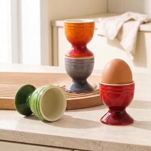 Może przedstawiać: Zestaw czterech ceramicznych kieliszków do jajek w kolorach pomarańczowym, szarym, zielonym i czerwonym. Czerwony kieliszek zawiera brązowe jajko. Pomarańczowy i szary kieliszek są ustawione jeden na drugim, a zielony leży na boku. Każdy ma szeroką podstawę.
