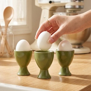 May include: Three sage green ceramic egg cups with a speckled finish, each holding a white egg. The cups have a pedestal base and a rounded bowl shape. The set is arranged on a wooden surface, with a cream-colored stand mixer in the background.