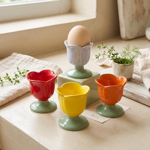Może przedstawiać: Zestaw czterech ceramicznych kieliszków do jajek w kształcie tulipanów, z płatkami w kolorze czerwonym, żółtym, pomarańczowym i jasnoniebieskim oraz szałwiowo-zielonymi łodygami i podstawami. Jasnoniebieski kieliszek zawiera brązowe jajko.