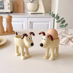 May include: Two cream-colored ceramic egg cups shaped like dogs with brown ears and black noses. One cup is empty, and the other holds a single brown egg. The figures feature four legs, a small tail, and wide-set eyes, designed to hold an egg securely.