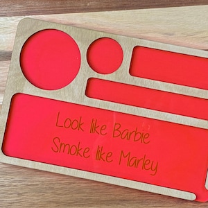 May include: A wooden rolling tray with a red, felt-like lining. The tray has multiple compartments and a cutout for a grinder. The tray has the text "Look like Barbie Smoke like Marley" written on it.