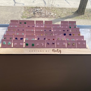 May include: A wooden display stand with a variety of colorful earrings. The earrings are arranged in rows on small cards with the text "Designs by Doty".
