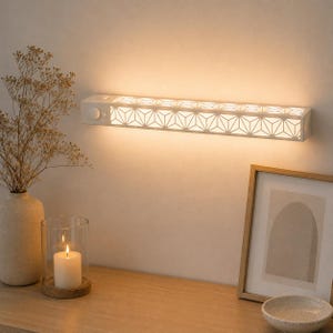 May include: A rectangular white wall-mounted light fixture featuring a geometric triangular lattice pattern that emits a warm glow. The fixture includes a circular sensor and a small switch on the left side, providing soft illumination.