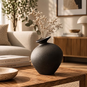 Può includere: Un vaso in ceramica nero opaco con un corpo arrotondato e bulboso e un collo stretto e svasato. Il vaso contiene una delicata composizione di fiori bianchi essiccati e steli di eucalipto verdi, creando un contrasto con la superficie scura e ruvida.