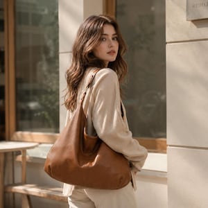 May include: A large, slouchy hobo-style shoulder bag crafted from smooth, tan-colored leather. The bag features a single wide strap, a spacious main compartment with a curved top opening, and clean, minimalist stitching along the exterior seams.