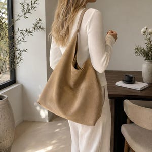 Puede incluir: Un bolso de hombro grande estilo hobo de color tostado con una textura suave y mate. El bolso cuenta con una correa ancha integrada que descansa sobre el hombro, creando una silueta holgada y desestructurada que cae a la altura de la cadera.