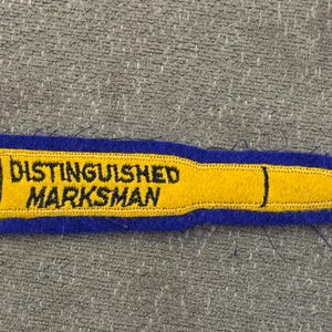 Può includere: Una toppa ricamata a forma di proiettile con un centro giallo, il testo nero "DISTINGUISHED MARKSMAN" e un bordo blu. Il design include dettagli a linee nere che rappresentano il bossolo e la punta del proiettile su una base in feltro.