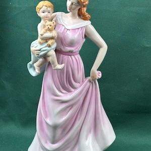 May include: A porcelain figurine depicting a person with reddish-brown hair wearing a flowing pink dress, holding a small child who is wrapped in a light blue cloth and clutching a tan teddy bear. The figures have painted facial features.