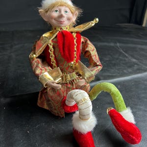 May include: A decorative elf doll featuring a pointed hat with white faux fur trim, a red and gold patterned tunic with gold metallic accents, and a red tie. The doll has one white striped leg and one green leg, both ending in red velvet shoes with bells.