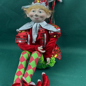 May include: A decorative elf doll featuring a red velvet outfit with silver sequin accents, a pointed hat with silver bells, and green and red diamond-patterned leggings. The doll has light-colored hair, pointed ears, and curled red velvet shoes.