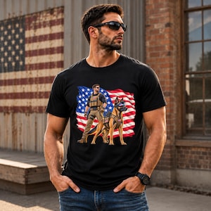 May include: A black t-shirt featuring a graphic print of a person in tactical gear standing next to a German Shepherd dog, both positioned in front of a waving American flag. The person wears sunglasses and a vest, while the dog wears a harness.