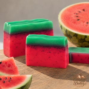 Peut inclure: Deux pains de savon rectangulaires conçus pour ressembler à des tranches de pastèque, avec une base rouge vif parsemée de petits points noirs et une couche supérieure verte. Une demi-pastèque et deux petits quartiers sont visibles.