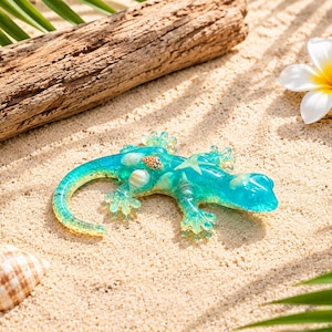 Peut inclure: Une figurine de gecko en résine turquoise translucide, ornée de minuscules coquillages et d'une petite étoile de mer incrustés sur le dos. L'ornement en forme de lézard présente un dégradé de tons bleus et jaunes le long de sa queue courbée.