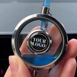 May include: A circular silver-toned metal keychain featuring a floating inner disc with the text YOUR LOGO printed in white on a black background. The outer ring contains a clear liquid with a blue tint that moves within the frame.