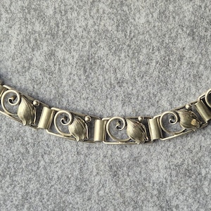 Op de afbeelding: Een zilverkleurige metalen armband bestaande uit vijf herhalende rechthoekige schakels. Elke schakel is voorzien van een gestileerd bloemknopmotief met een gebogen krul-element en een klein bolvormig accent, verbonden door gladde metalen staven.