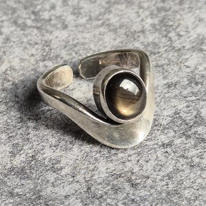 May include: A silver-toned open-band ring featuring a smooth, polished finish and a single, round, dark brown cabochon gemstone set in a circular bezel. The band has a curved, asymmetrical design that tapers towards the open ends at the back.
