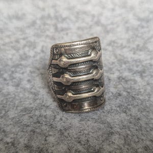 Op de afbeelding: Een brede zilverkleurige metalen ring met een getextureerd oppervlak en drie horizontale verhoogde balken in het midden. De ring heeft een donkere geoxideerde afwerking in de verdiepte delen, wat de ingewikkelde gegraveerde patronen langs de randen benadrukt.