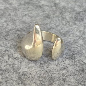 Puede incluir: Un anillo abierto de tono plateado con un acabado liso y pulido de diseño escultórico asimétrico. La banda está dividida, mostrando dos formas distintas, redondeadas y orgánicas que se curvan hacia arriba, creando una superficie metálica fluida y reflectante.