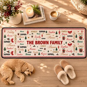 Puede incluir: Una alfombra rectangular de fondo crema con un patr&oacute;n navide&ntilde;o repetitivo de lazos rojos, &aacute;rboles verdes y cajas de regalo. El texto THE BROWN FAMILY est&aacute; centrado, rodeado por los nombres Emily, Sophia y Anna en diversas tipograf&iacute;as.