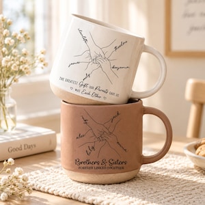 Peut inclure: Un mug en céramique blanc empilé sur un mug couleur terre cuite. Le mug blanc présente un dessin au trait de mains jointes avec les noms Charlotte, Theodore, Benjamin, Mely et James, et le texte THE GREATEST GIFT OUR PARENTS GAVE US WAS EACH OTHER. Le mug terre cuite montre des mains jointes avec les noms Ruby, Jack, Emma, Lily et Alex, et le texte BROTHERS & SISTERS FOREVER LINKED TOGETHER.